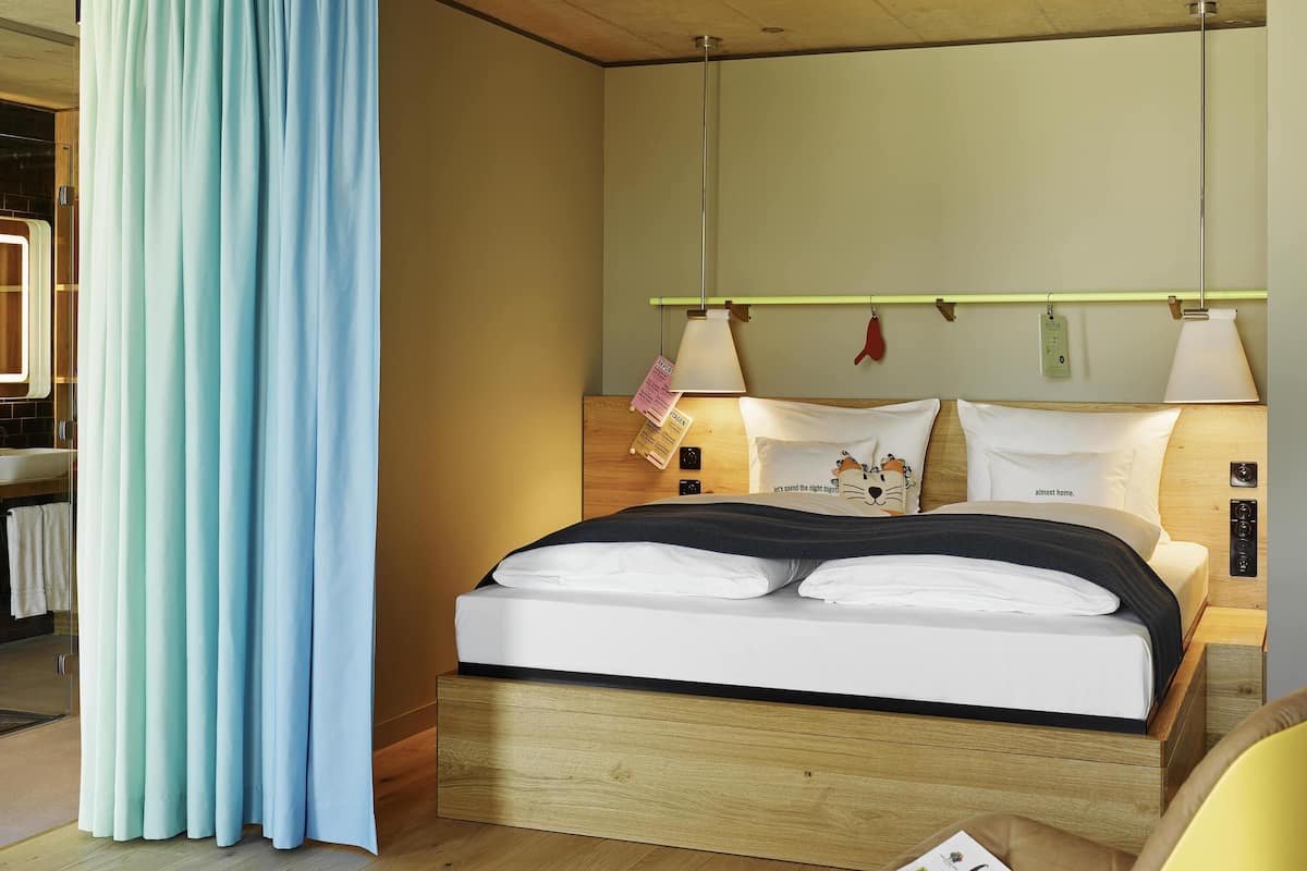 A neatly made bed with a headboard, a bedside table with a lamp, and a window with curtains.