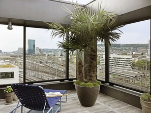 A modern room with a large window, a potted plant, a blue chair, and a newspaper. - 25hours Hotel Zurich Langstrasse (Zürich)