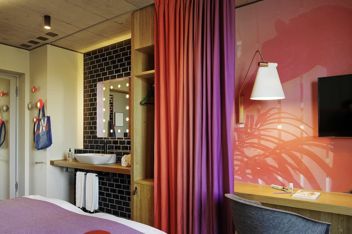 A modern hotel room with a bed, a desk, a chair, a vanity with a mirror, and a TV.