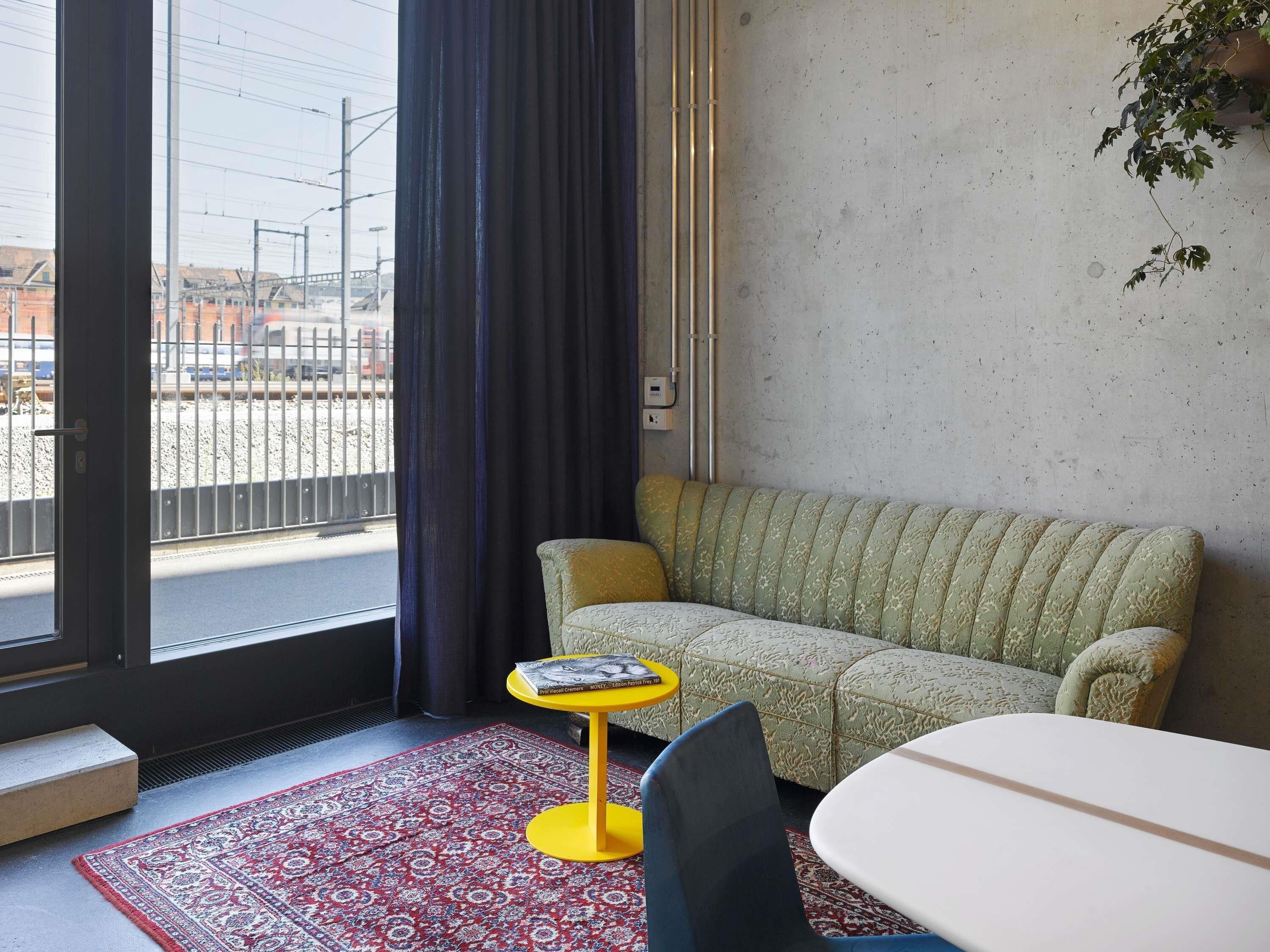 A room with a sofa, a small round table with a book, a chair, a red patterned rug, and a view of the outside through a window with a curtain.