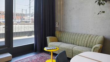 A room with a sofa, a small round table with a book, a chair, a red patterned rug, and a view of the outside through a window with a curtain.