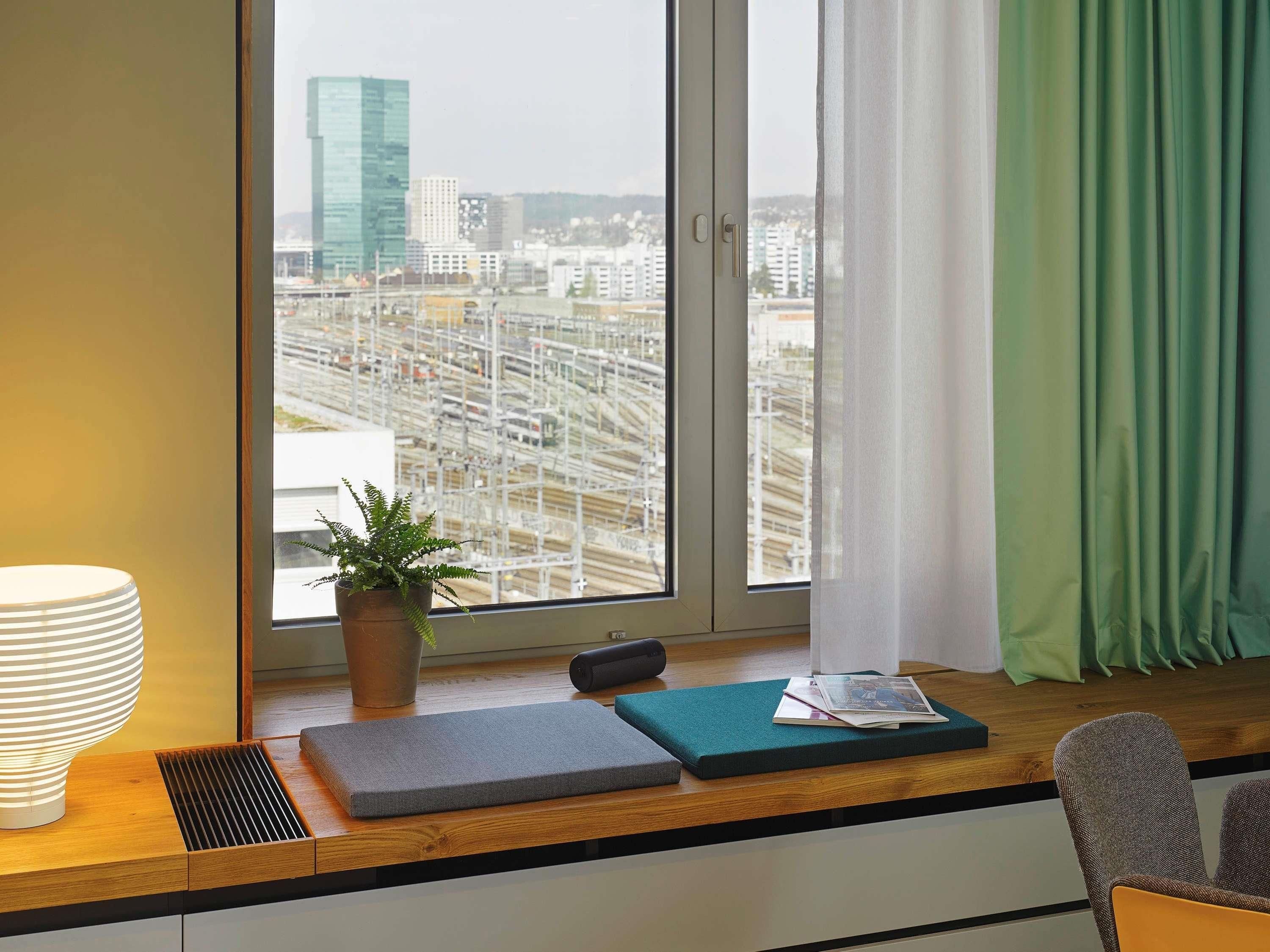 A room with a large window, a potted plant, a lamp, and a cushion on a wooden desk.