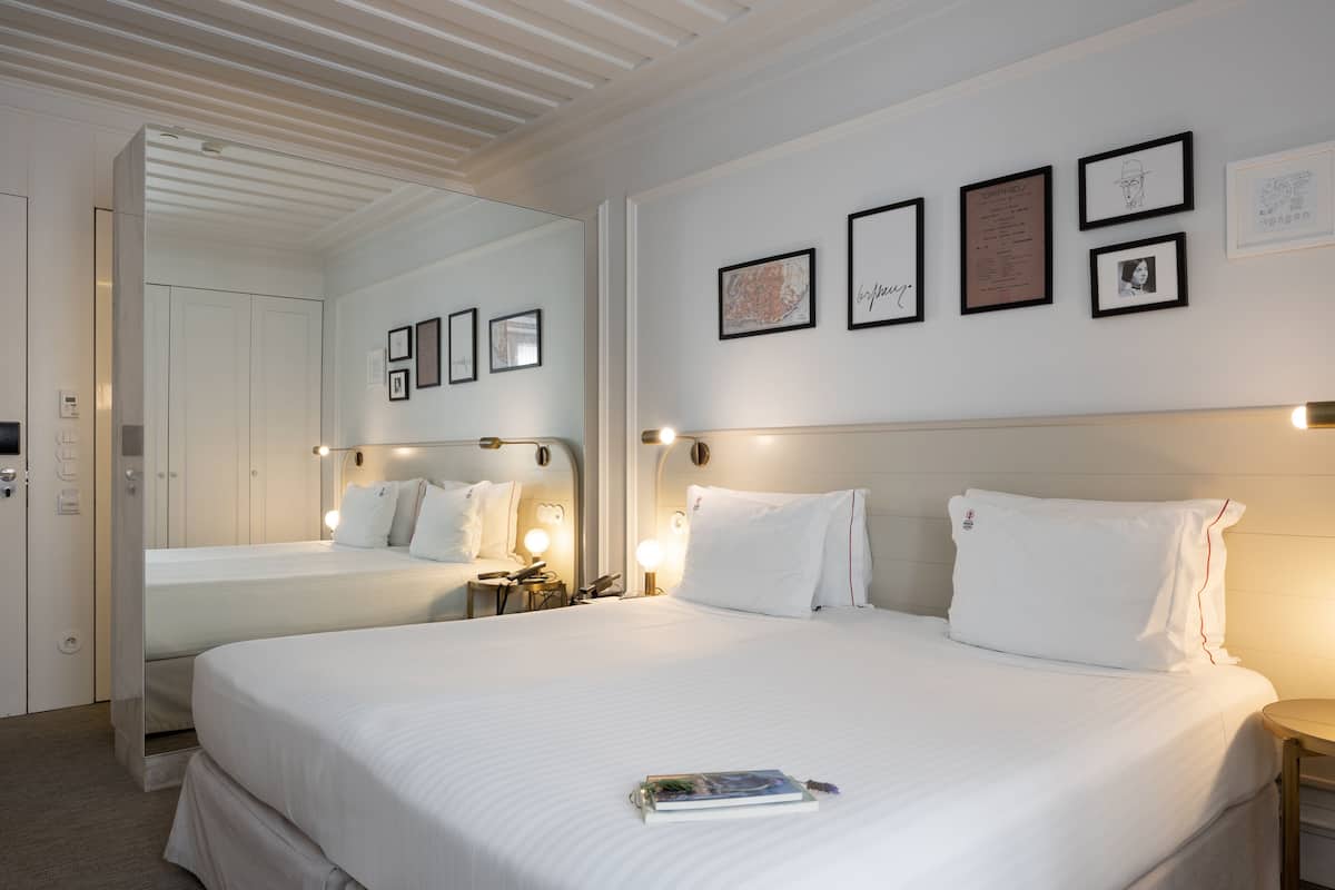 Une chambre d’hôtel avec deux lits, un grand miroir et des œuvres d’art encadrées accrochées au mur.