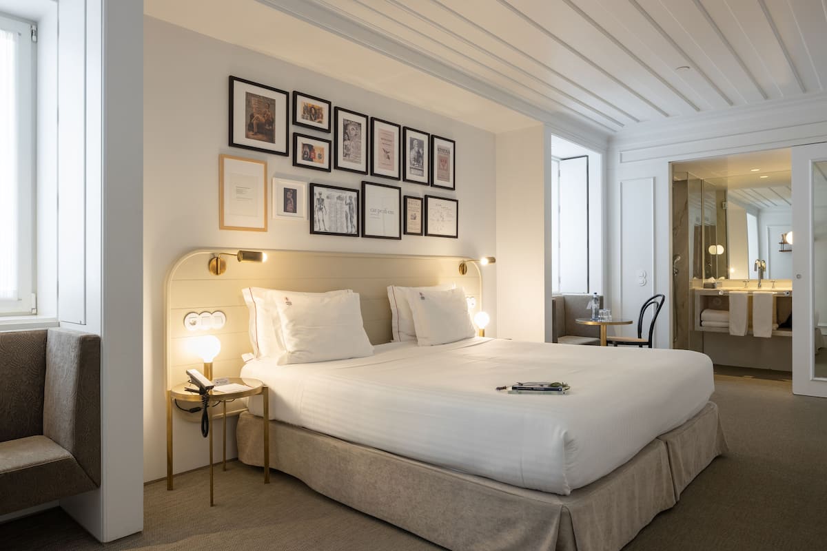 Une chambre d’hôtel bien aménagée, avec un grand lit, des tables de chevet dotées de lampes, un mur orné de tableaux encadrés et une salle de bain visible à travers une porte ouverte.