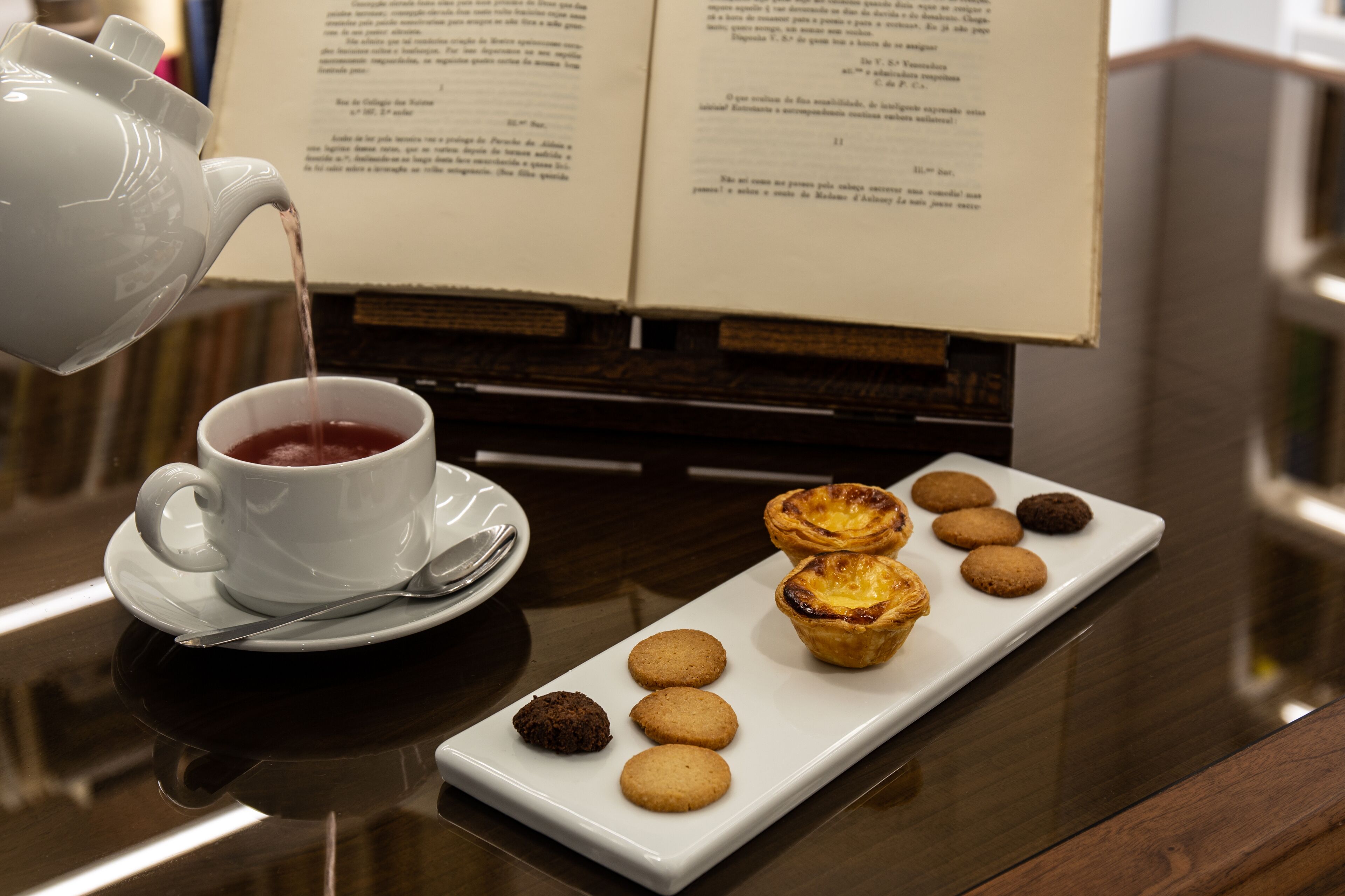 A table with a white cup, saucer, and spoon, a plate with cookies and a tart, and a book with text.