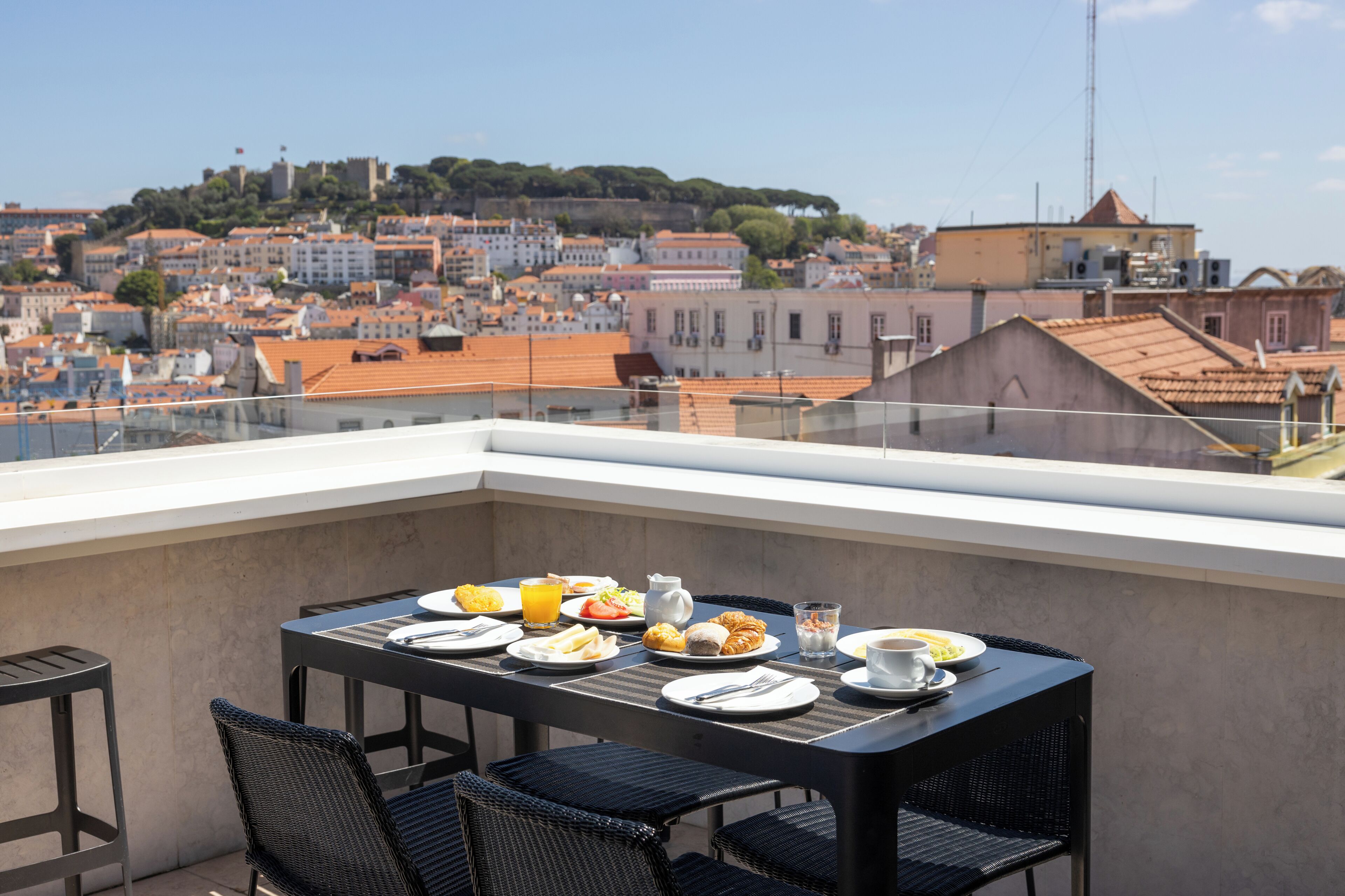 Un balcon avec une table dressée pour le petit-déjeuner, d’où l’on peut admirer un panorama sur la ville, avec ses bâtiments et une colline.