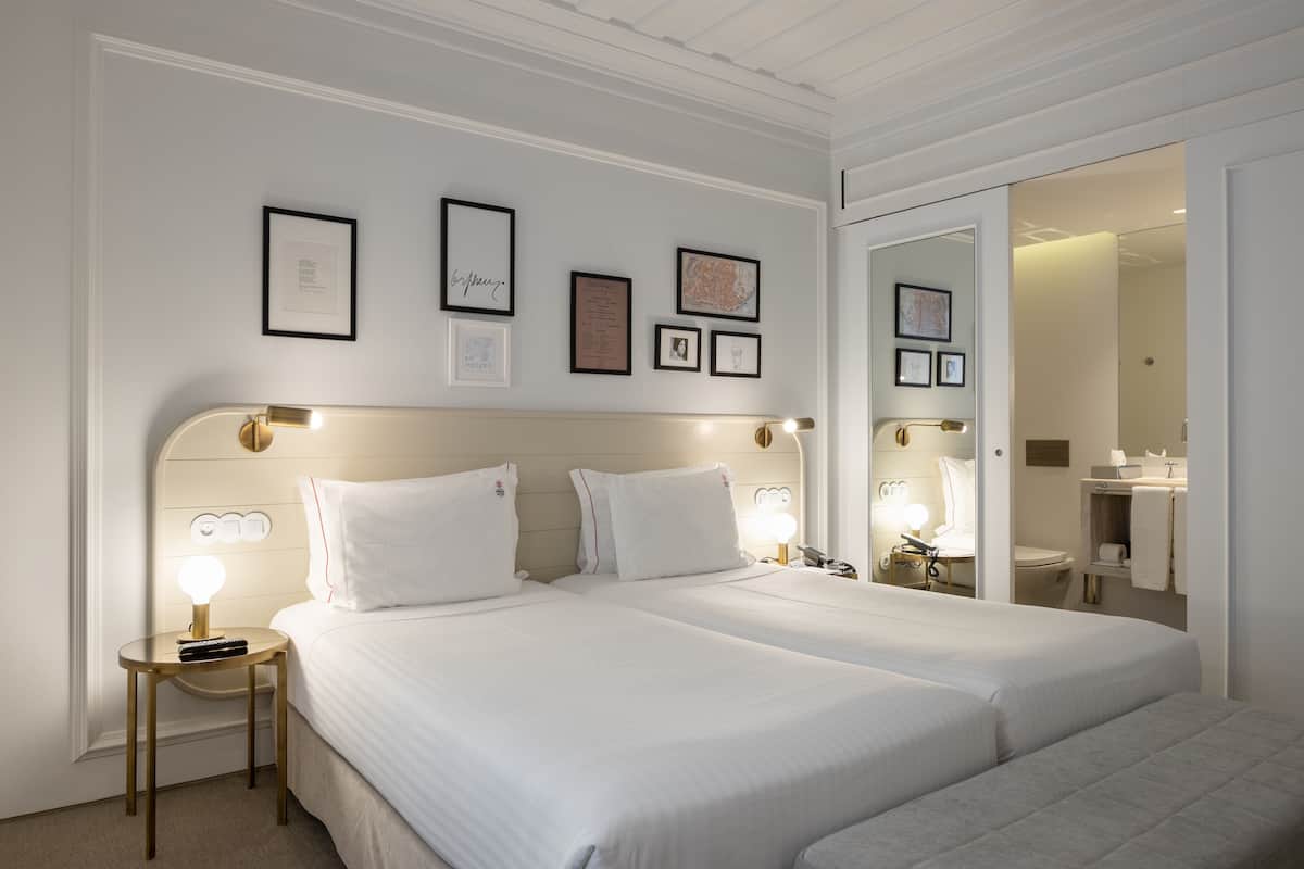 Une chambre d’hôtel avec deux lits, une table de chevet avec une lampe, des tableaux encadrés au mur et une salle de bain visible à travers une porte ouverte.