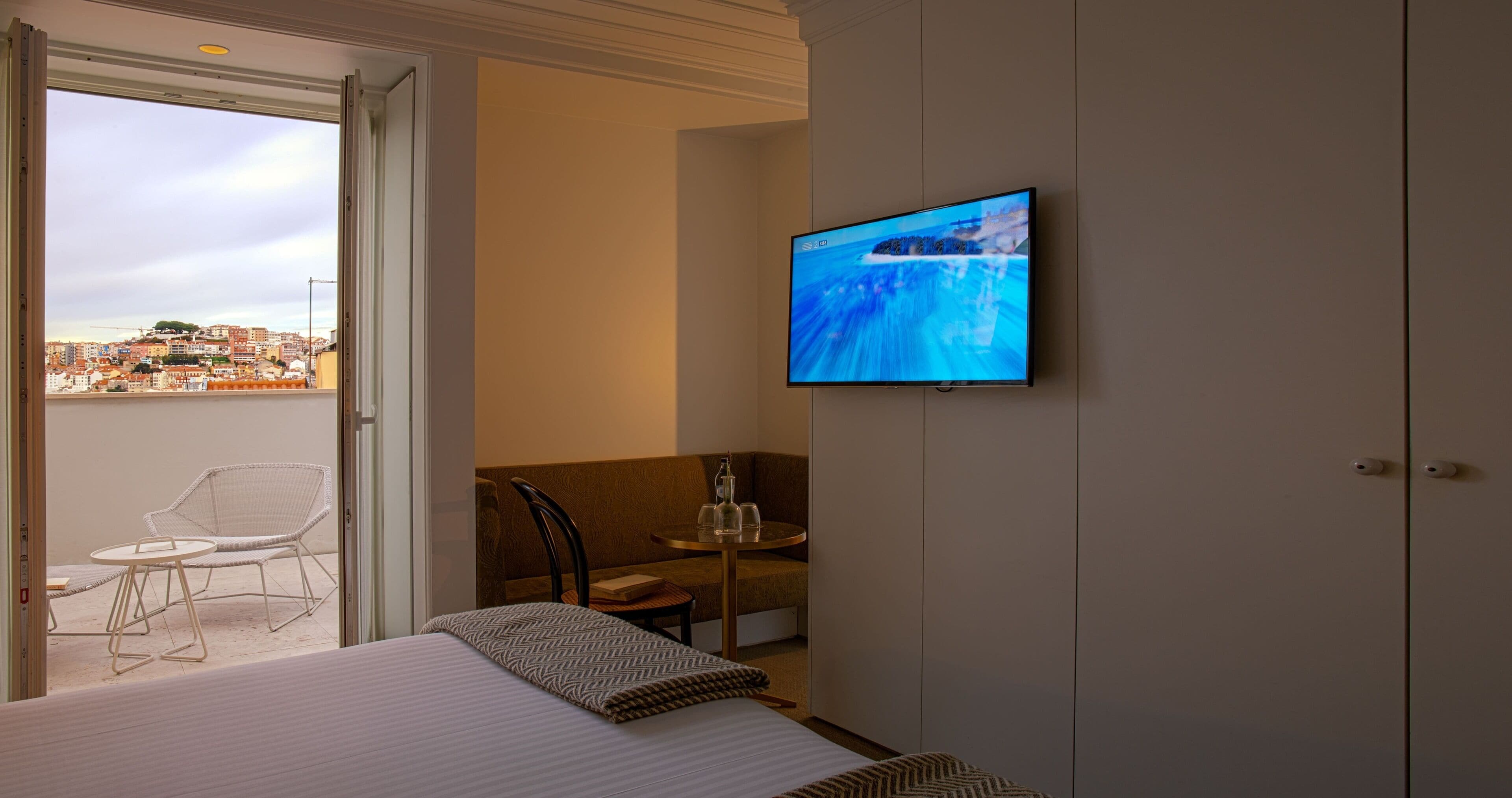 Een hotelkamer met een bed, een aan de muur bevestigde televisie, een balkon met een stoel en tafel, en uitzicht op de stad.