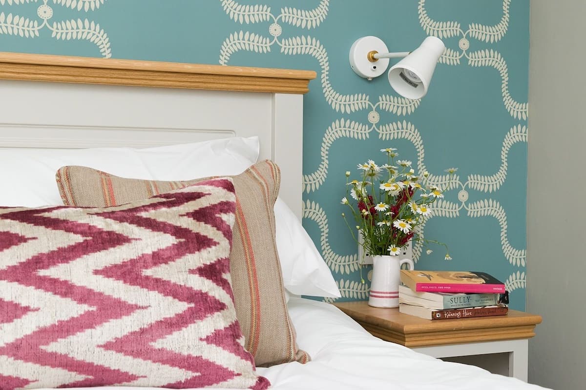 A neatly made bed with a patterned throw, a bedside table with a vase of flowers, and a wall-mounted lamp.