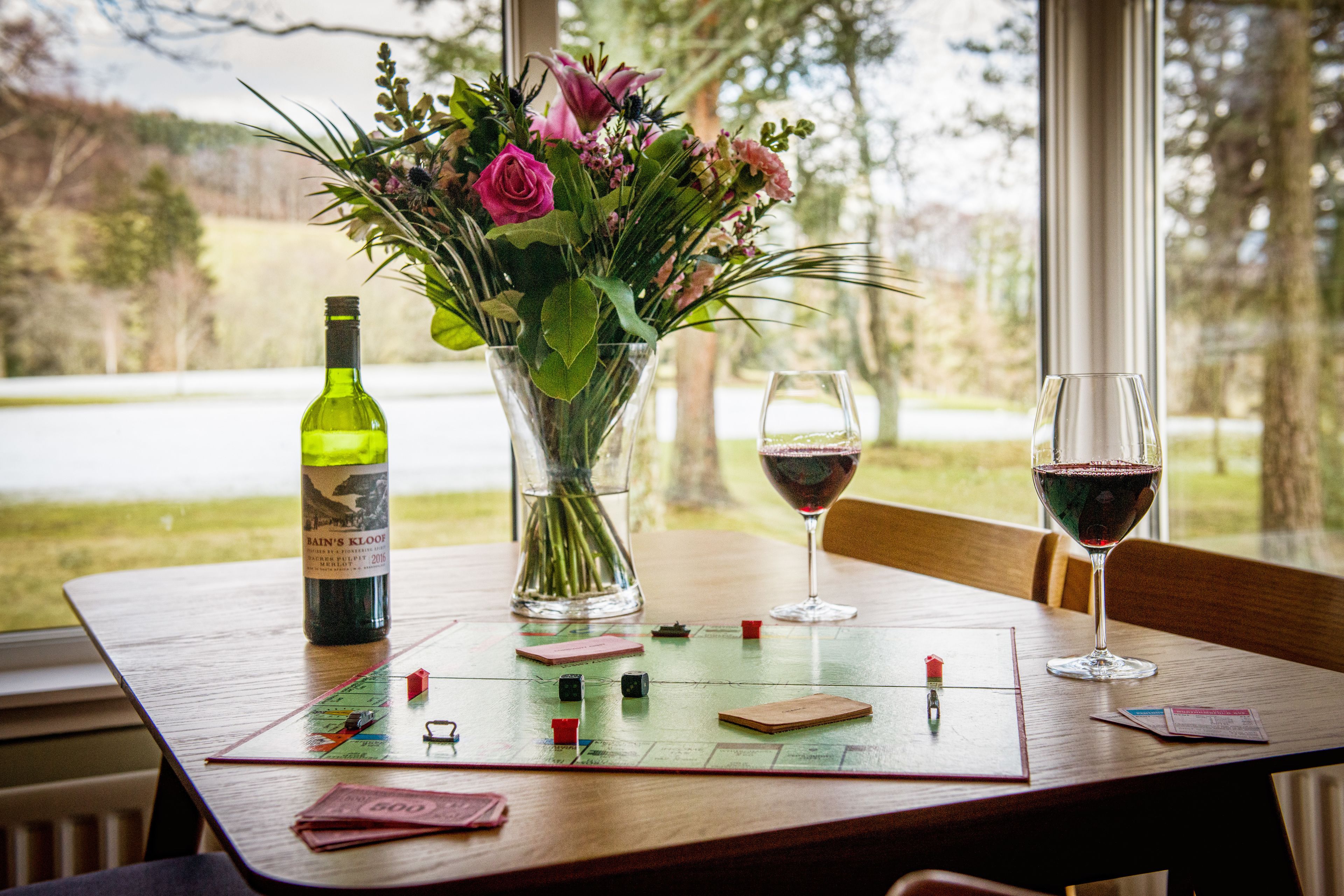 A wooden table with a bottle of wine, a vase with flowers, two wine glasses, and playing cards.