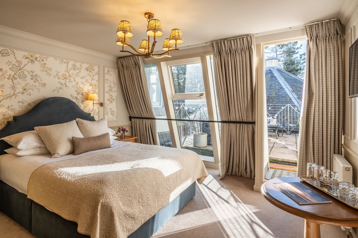 A bedroom with a large bed, a chandelier, a view of the outside, and a small table with glasses.