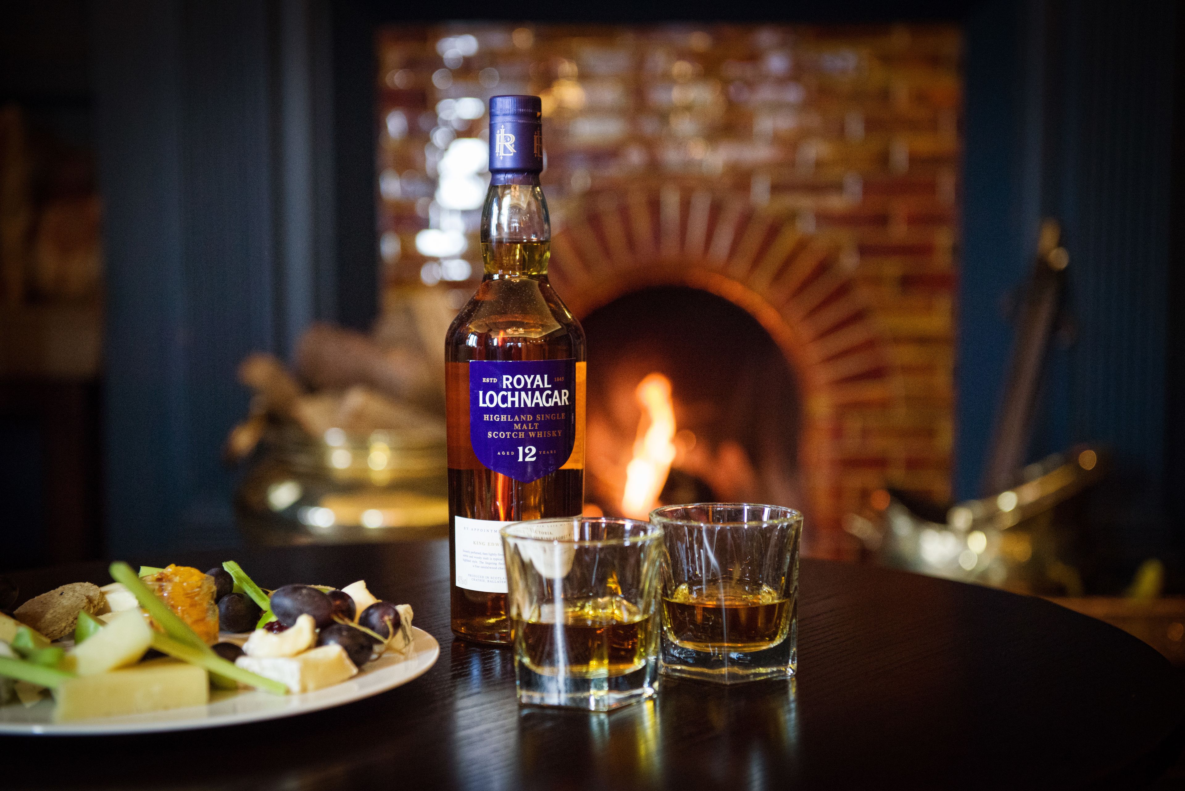 A bottle of Royal Lochnaggar whisky, two shot glasses, and a plate of cheese and fruits on a table with a brick fireplace in the background.