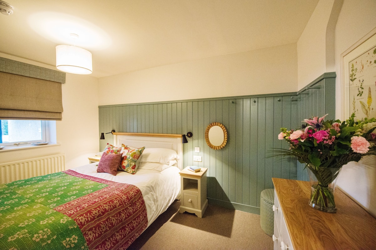 A bedroom with a bed, bedside table, nightstand, and a vase of flowers.