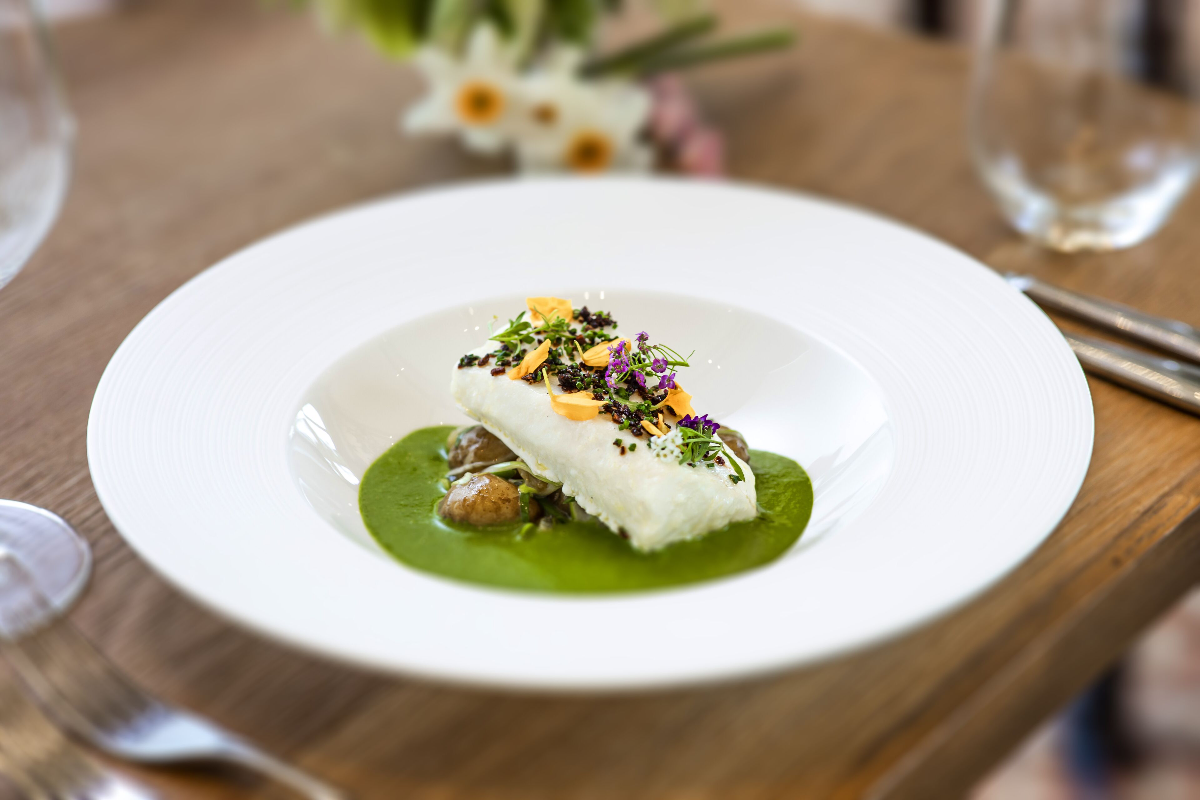Un plato blanco con un guiso de pescado, salsa verde y adornos sobre una mesa de madera.