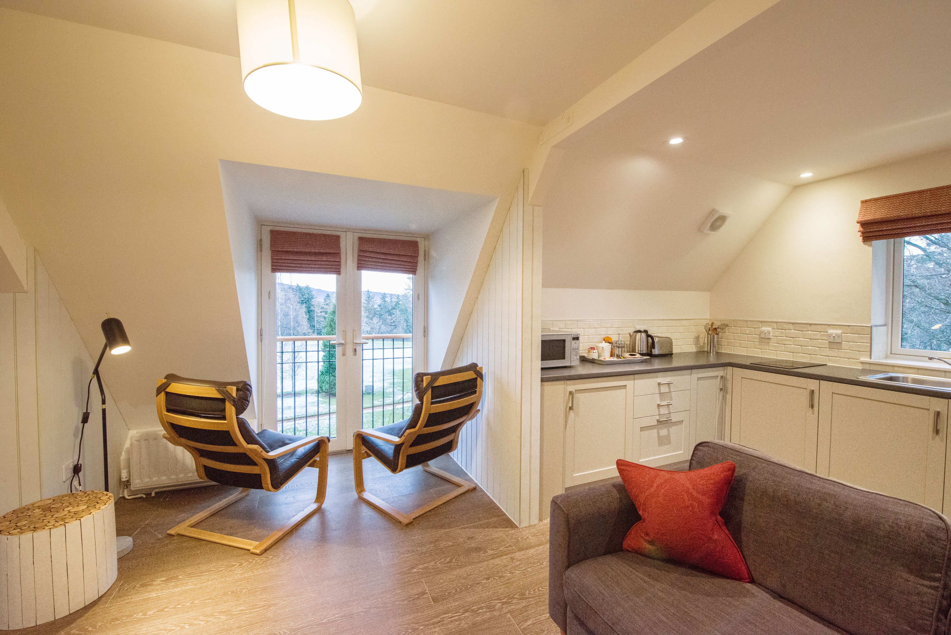 Een gezellige kamer met een bruine bank, twee gestreepte stoelen, een kleine keukenhoek en een raam met gordijnen.
