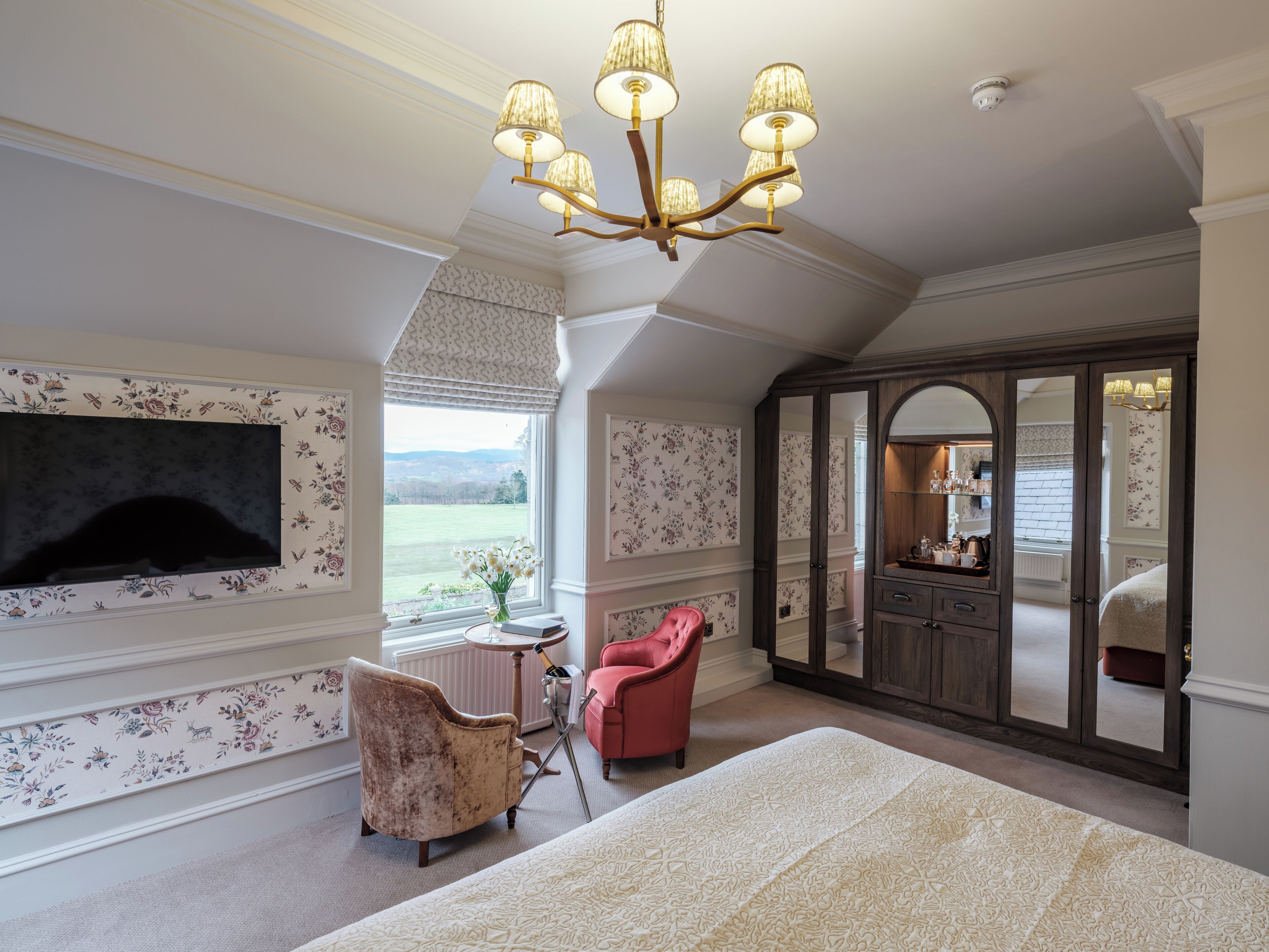 A bedroom with a large TV, a chandelier, a red chair, and a view of the outdoors.