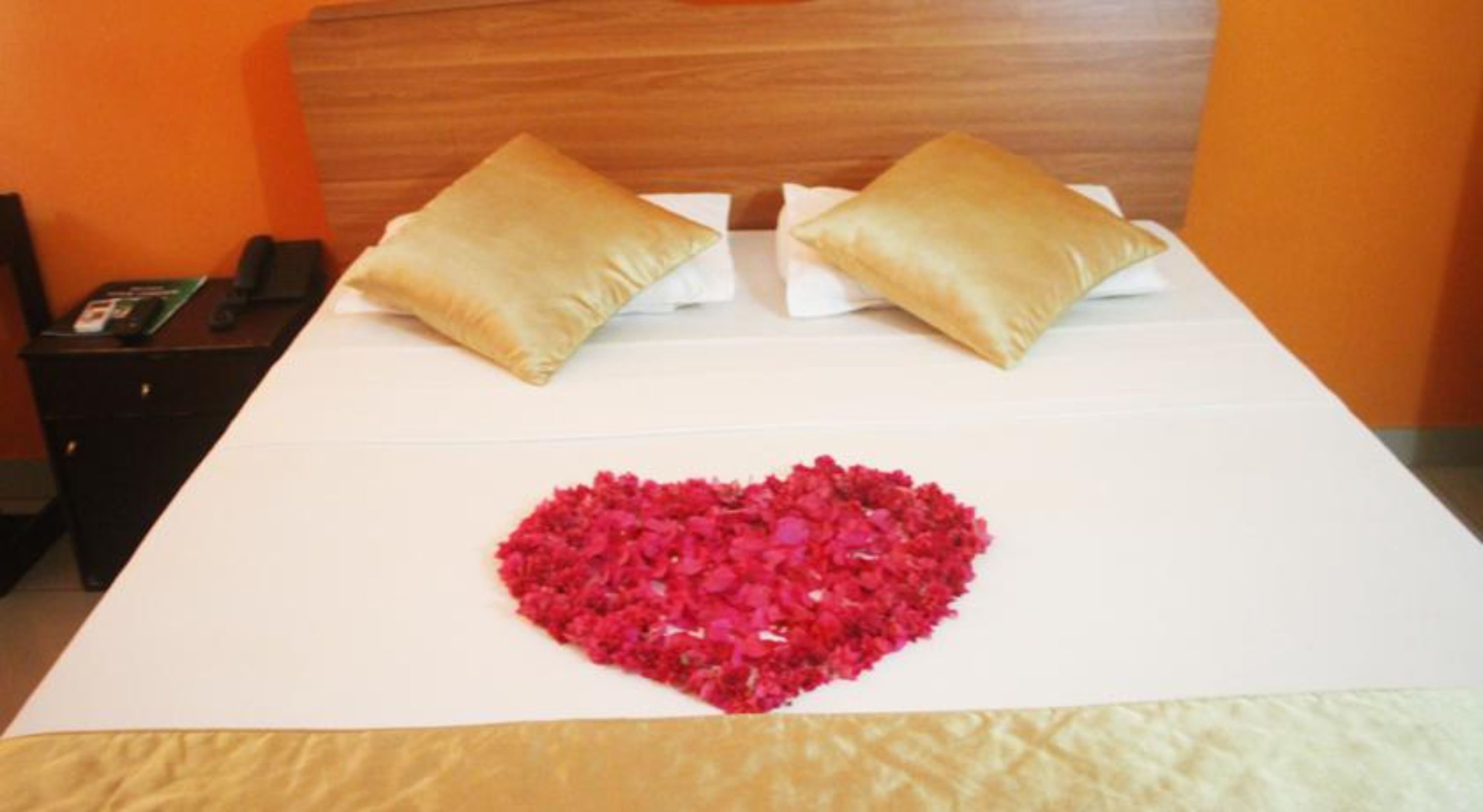A hotel room with a bed, two pillows, a bedside table with a phone, and a heart-shaped arrangement of rose petals on the bed.