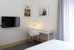 A hotel room with a white desk, a chair, a flat-screen TV, and two framed pictures on the wall.