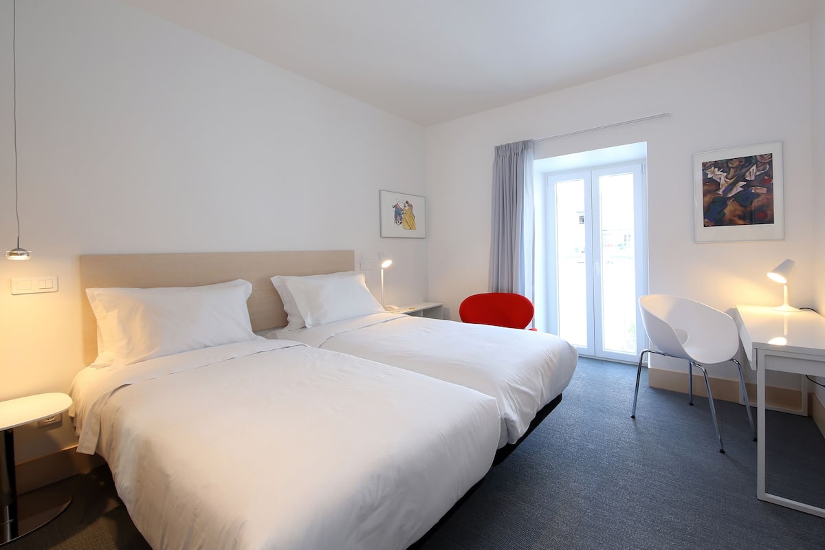 Una habitación de hotel con dos camas, un escritorio, una silla y una ventana con cortinas.