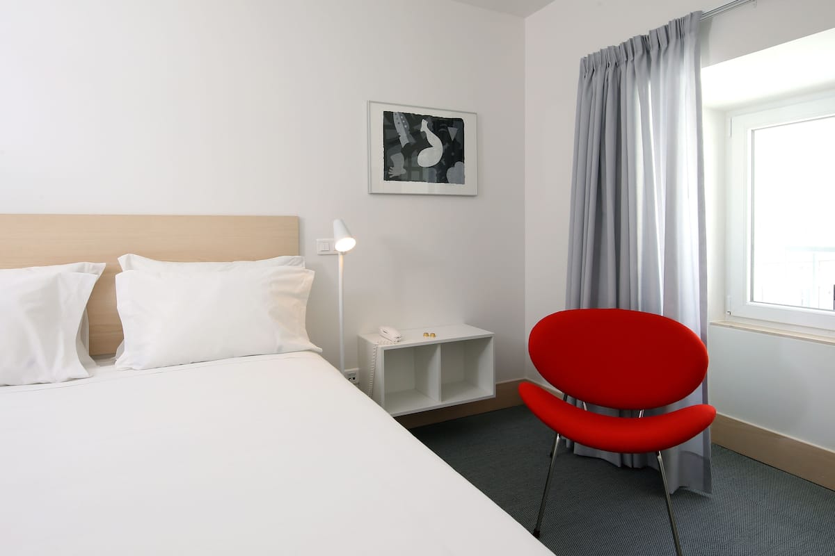 Una habitación de hotel con una cama, una silla roja, una mesita de noche con una lámpara, un teléfono y un cuadro en la pared.