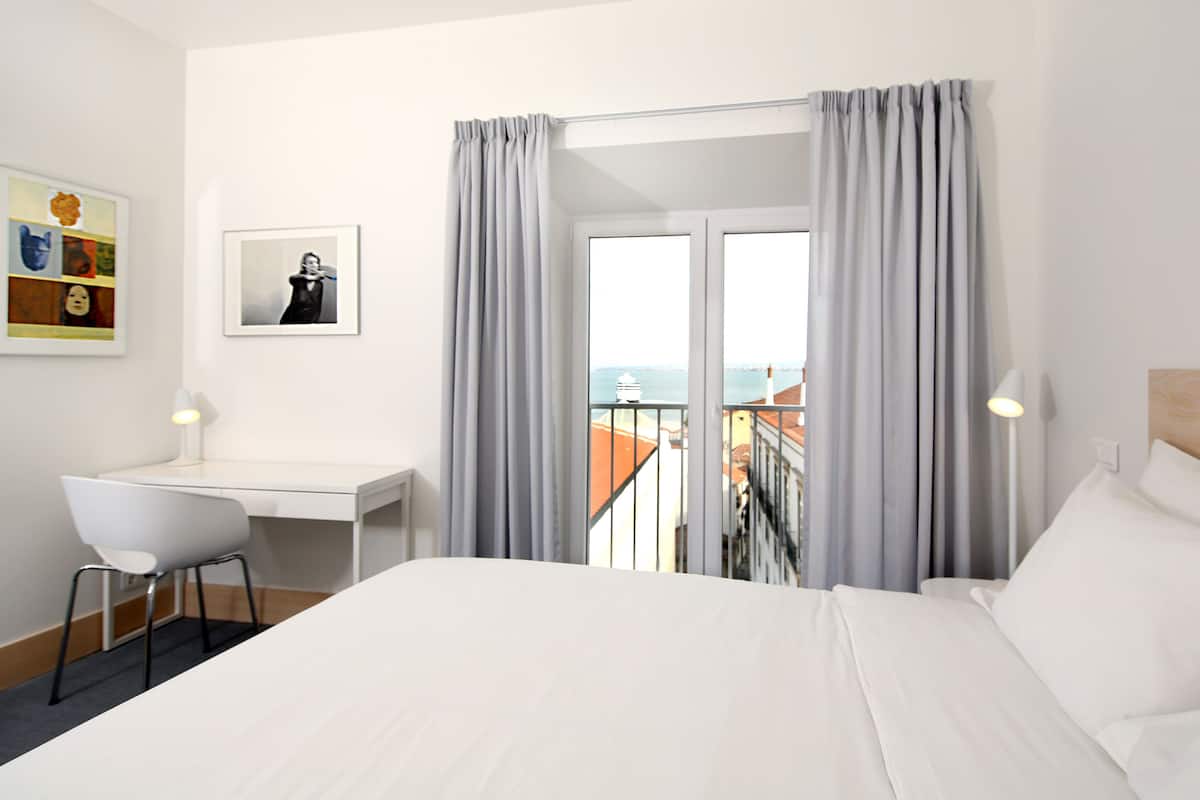 Habitación de hotel con cama, escritorio y una silla. Hay una ventana con cortinas, y un cuadro en la pared.