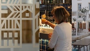 Een modern interieur met een bar, een persoon die een drankje bereidt en uitzicht op een gebouw met terrasplekken.