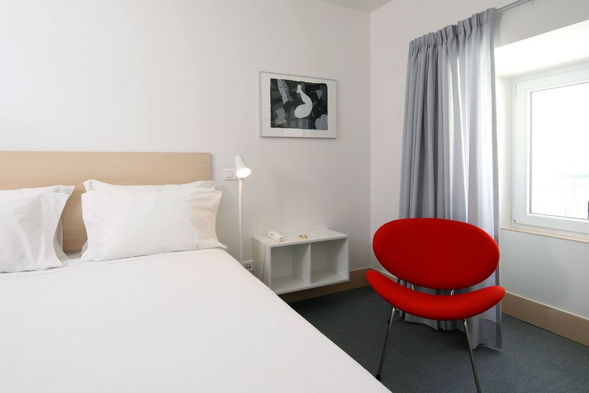 Una habitación de hotel con una cama, una silla roja, una mesita de noche con lámpara, un teléfono y una obra de arte enmarcada en la pared.