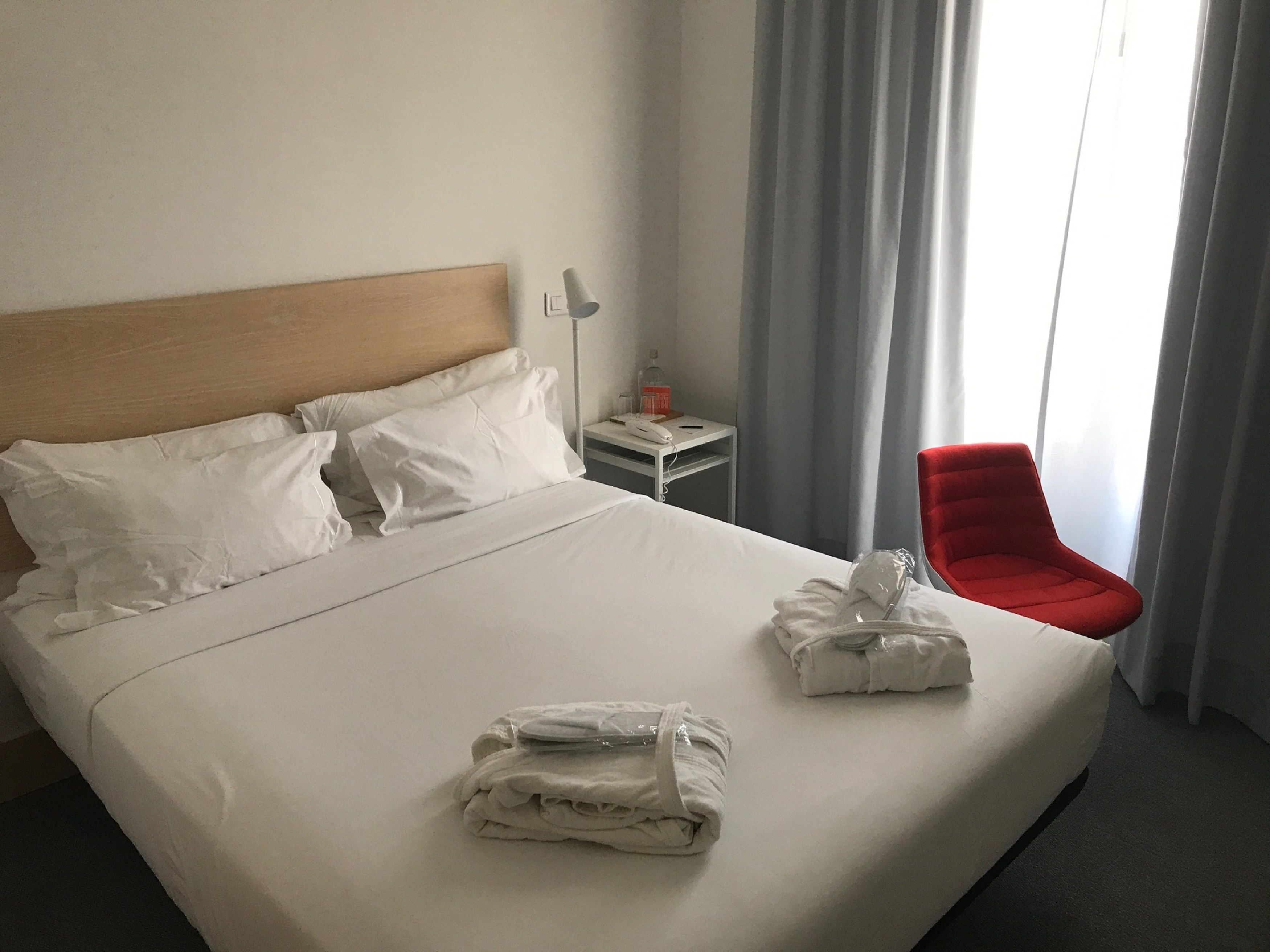 A hotel room with a large bed, white linens, a red chair, and a nightstand with a lamp and phone.