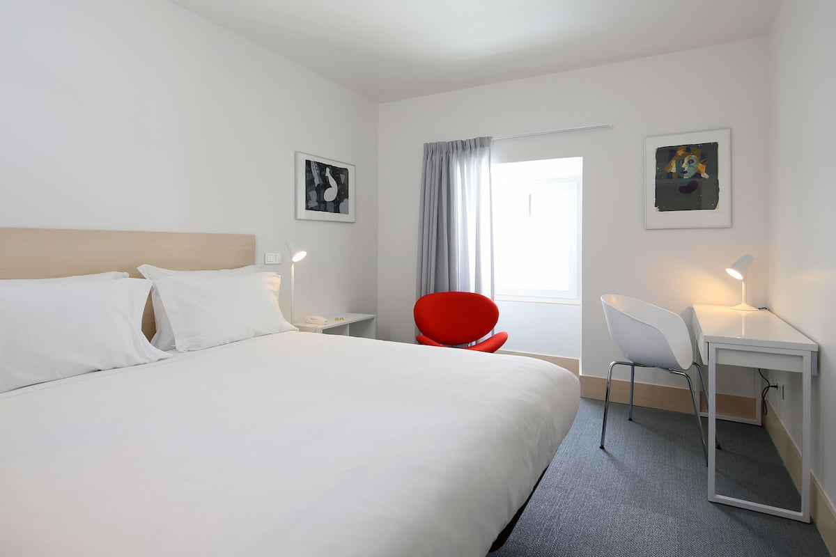 Una habitación de hotel con una cama grande, una silla roja, un escritorio y dos cuadros en las paredes.