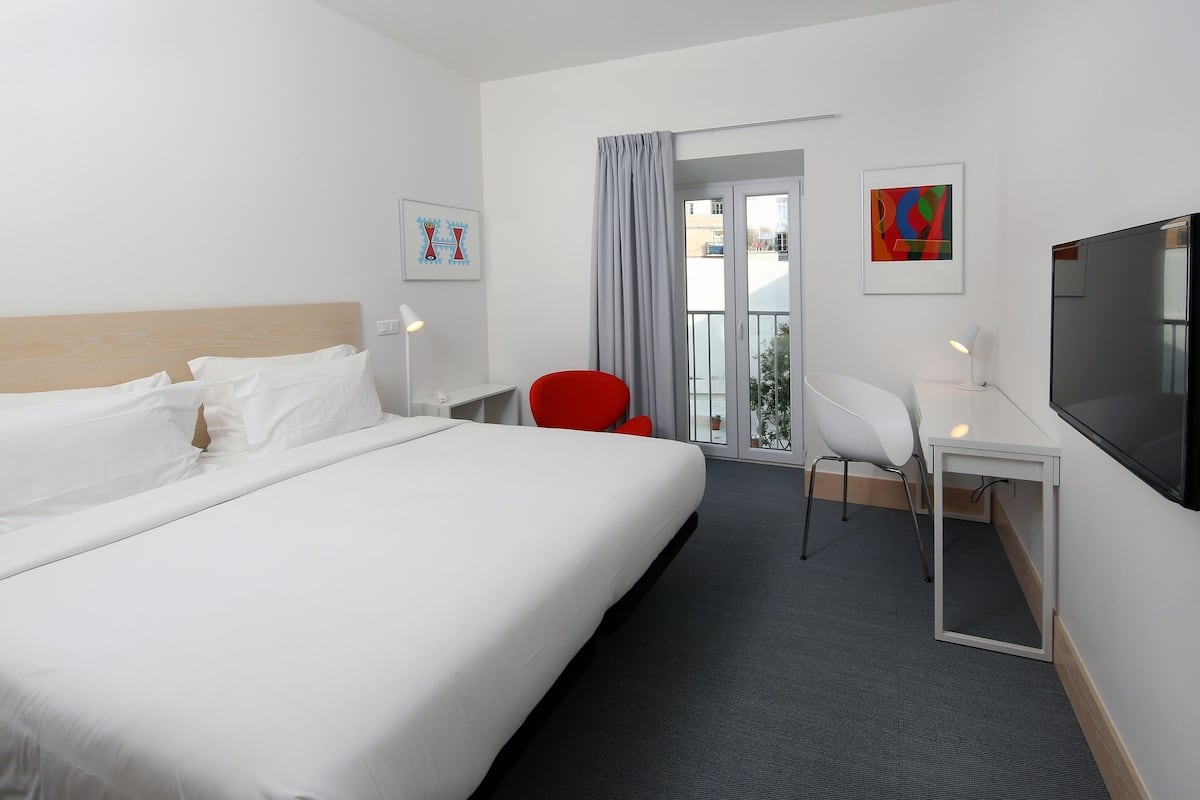Habitación de hotel con cama, escritorio, televisión, una silla roja y balcón con vistas.