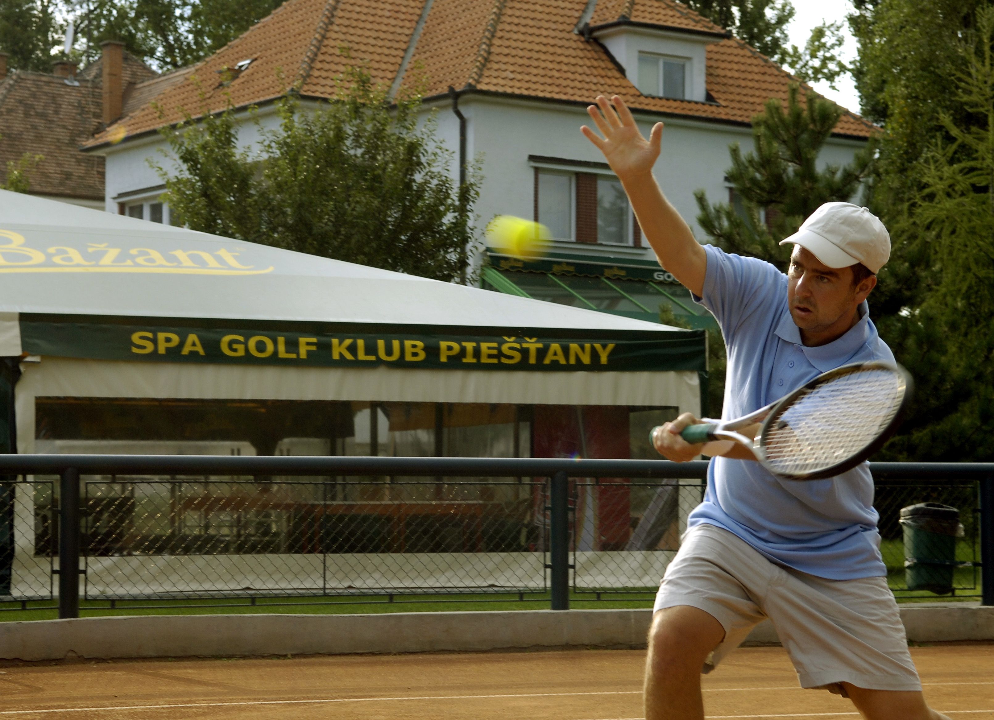 Un tenista en acción en una cancha de arcilla, con un edificio que dice "SPA GOLF KLUB PIESTANY" al fondo.