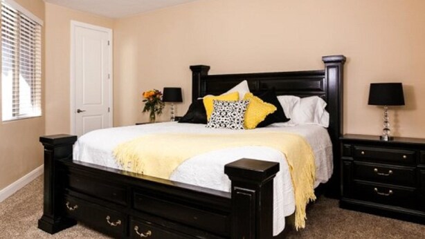 A bedroom with a black bed frame, white bedding, and yellow accents. There are two bedside tables with lamps, a door, and a window with blinds. - Zion Escapes (Washington)
