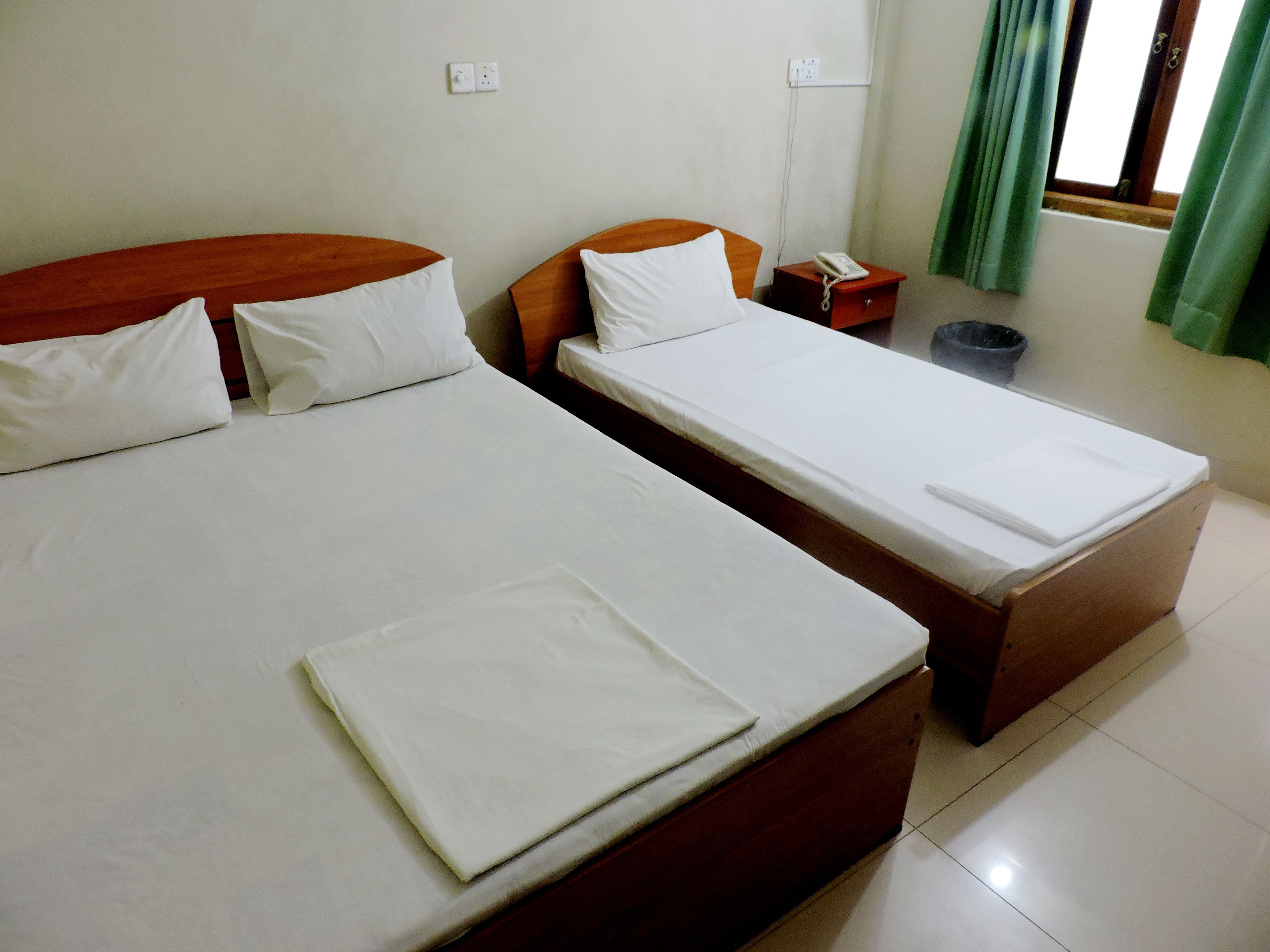 A hotel room with two beds, a wooden headboard, a bedside table with a phone, and a window with green curtains.