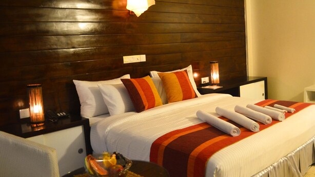 Habitación de hotel con cabecera de madera, ropa de cama blanca y una mesita de noche con lámpara y frutero.
