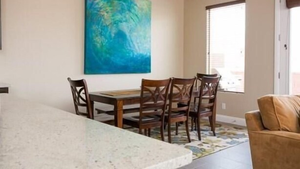 Un comedor con una mesa y sillas de madera, un cuadro abstracto grande en la pared y una ventana con persianas.
