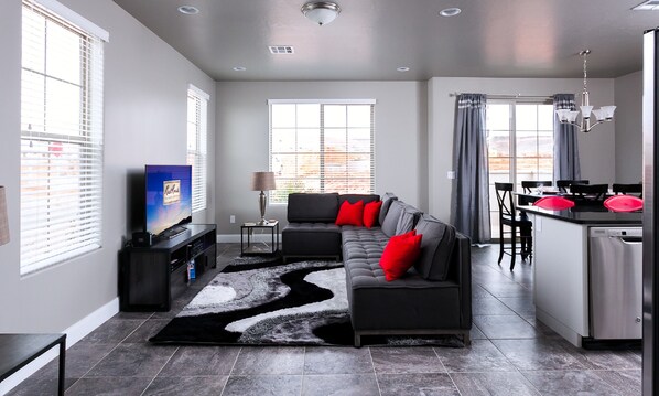 A modern living room with a sectional sofa, a flat-screen TV, and a kitchen area with a dining table and chairs.