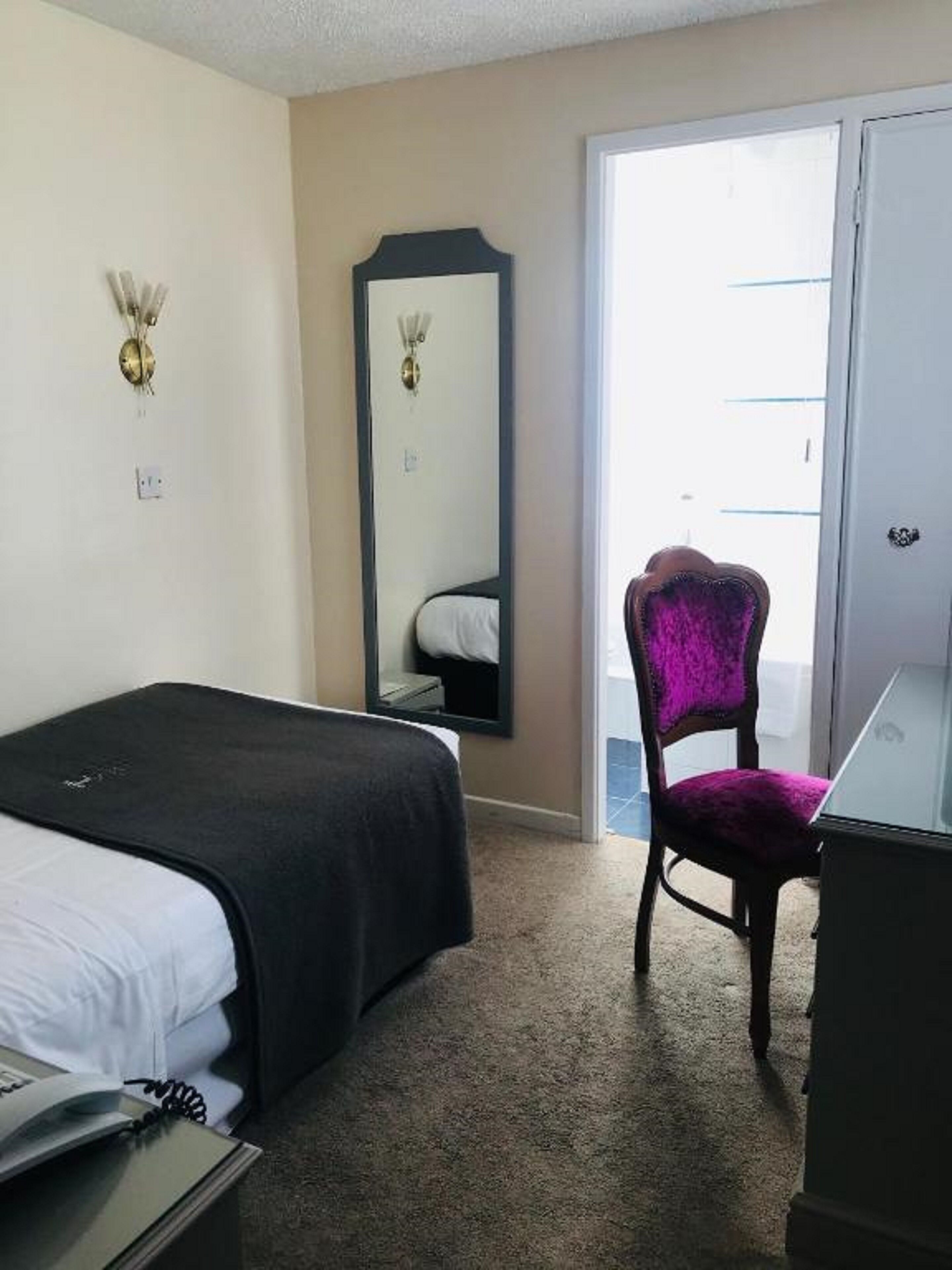A hotel room with a bed, a chair, a desk with a laptop, a mirror, and a door leading to a balcony.