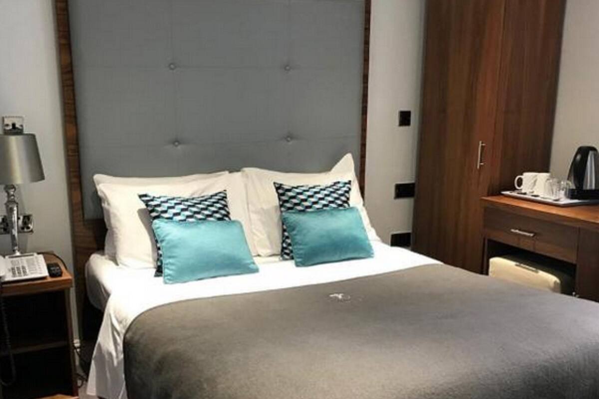 A neatly made bed with a grey blanket and two teal pillows, a wooden nightstand with a lamp, and a wooden wardrobe.