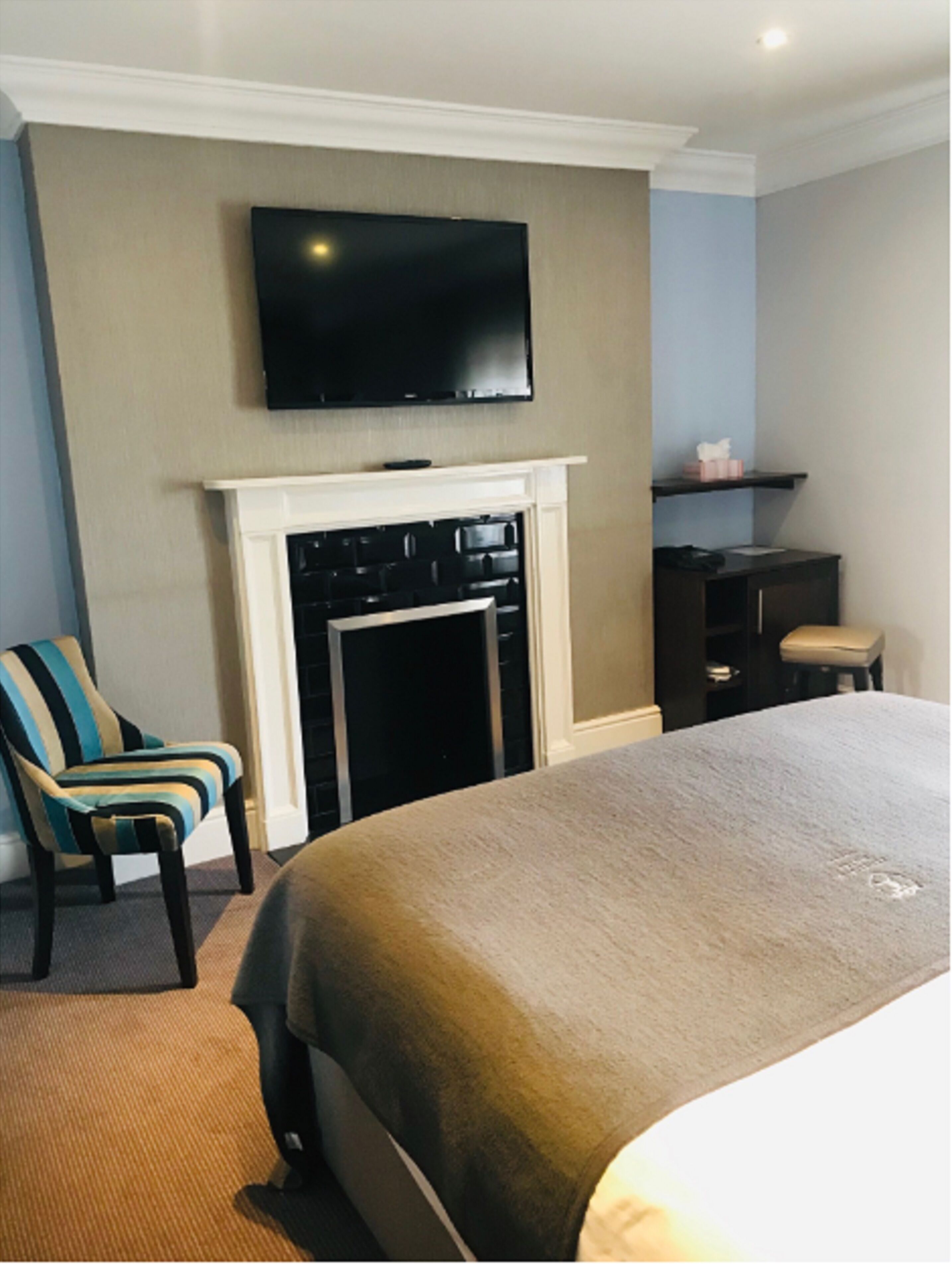 Een hotelkamer met een groot bed, een flatscreen televisie boven een open haard, een gestreept fauteuil en een klein bureau met stoel.