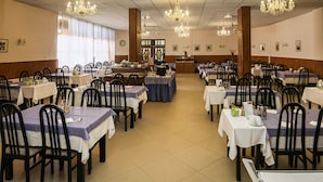 A spacious dining hall with multiple tables and chairs, chandeliers, and a clock on the wall. - Ensana Vila Trajan (Piestany)