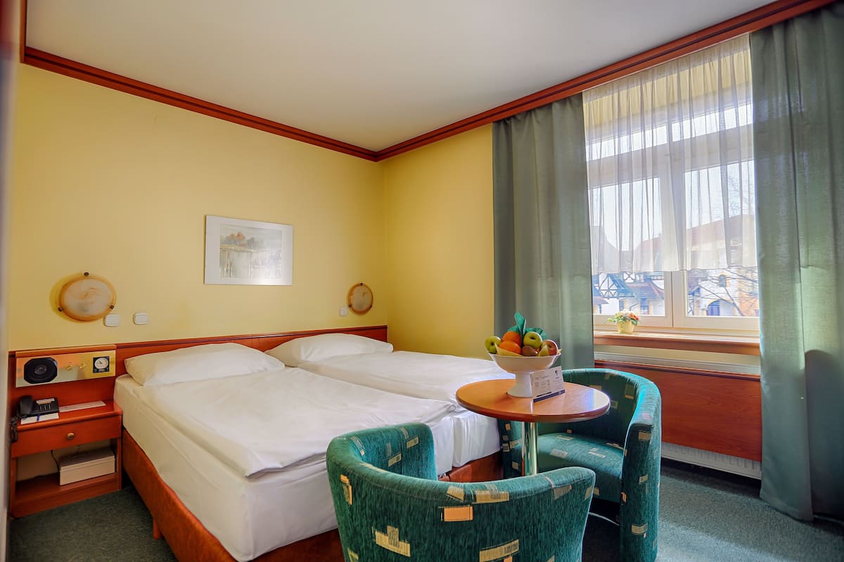 Ein Hotelzimmer mit zwei Betten, einem kleinen Tisch mit Früchten, einem Stuhl, einer Uhr und einem Fenster mit Vorhängen.
