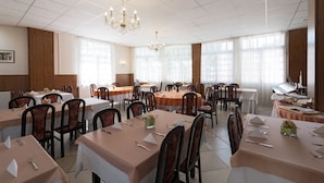A spacious dining area with multiple tables and chairs, large windows with sheer curtains, and a chandelier. - Ensana Vila Trajan (Piestany)