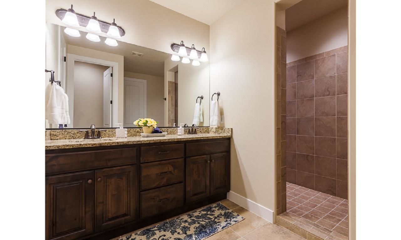 A bathroom with a dark wood vanity, a large mirror with lights, a shower with brown tiles, and a blue patterned rug.