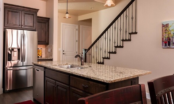 A kitchen with granite countertops, stainless steel appliances, dark wooden cabinets, and a black staircase.