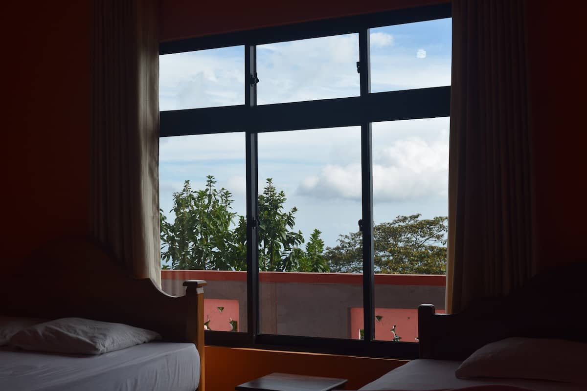 A room with two beds, a large window with a view of trees and clouds, and a balcony with a red railing.