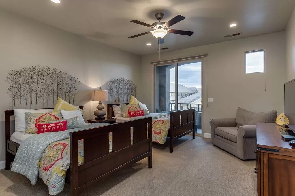 A bedroom with two beds, a ceiling fan, a window, and a view of a building.