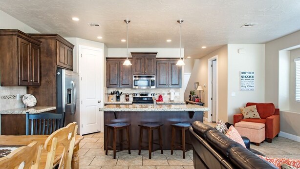 A modern kitchen with dark wood cabinets, a stainless steel refrigerator, a microwave, and a large island with bar stools. There is a red chair and a black leather couch in the adjacent living area. - Life is Good (Washington)