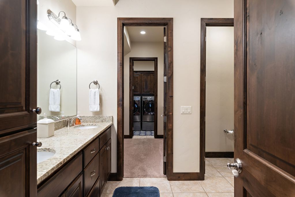 A bathroom with a double sink, vanity, and a mirror. There is a hallway leading to a laundry room with a washing machine and dryer.