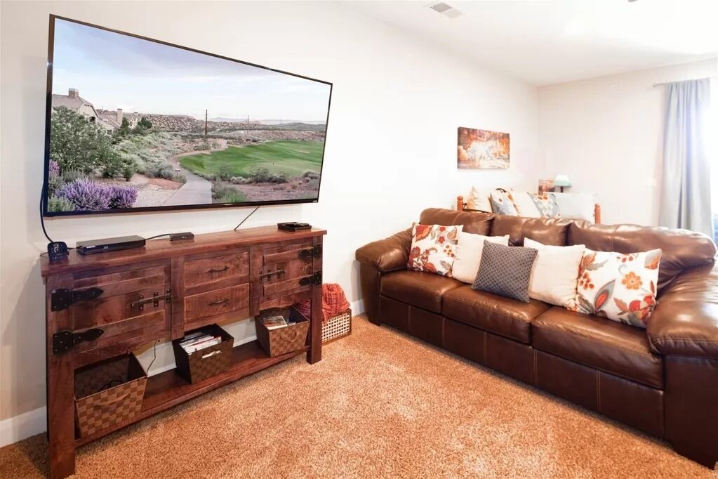 Een woonkamer met een flatscreen-televisie aan de muur, een bruine leren bank met gestreepte kussens en een houten dressoir met lades en planken.