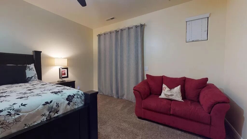 A bedroom with a bed, a red couch, a nightstand, a lamp, and a window with curtains.